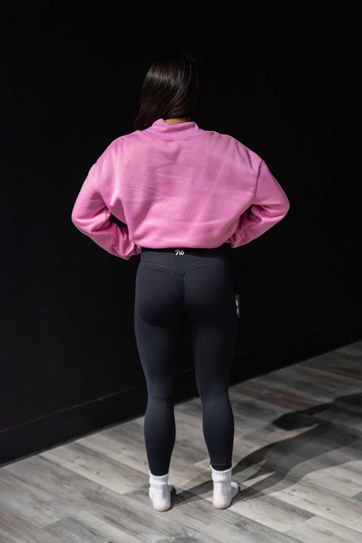 Person wearing a pink sweatshirt and black leggings standing against a dark wall.